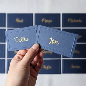 May include: Navy blue place cards with gold lettering. One card reads "Cecilia" and the other reads "Jon". Other cards with names are in the background. The cards have a rectangular shape with a decorative border.