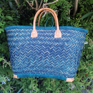 Könnte beinhalten: Ein gewebter blau-weißer Korbtasche mit braunen Ledergriffen und Akzenten. Die Tasche hat eine rechteckige Form mit einem gewebten Muster und einem türkisfarbenen Besatz. Die Griffe sind mit Lederstücken befestigt. Die Tasche steht vor einem Hintergrund aus grünem Laub.