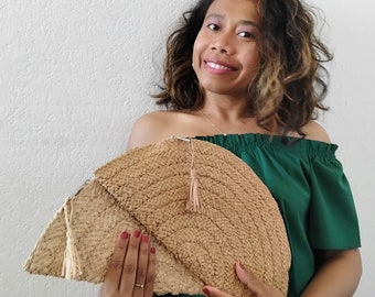 May include: A woman is holding a large, woven, light brown clutch with a tassel. The clutch is shaped like a half-circle and has a woven pattern. The woman is wearing a green off-the-shoulder top.