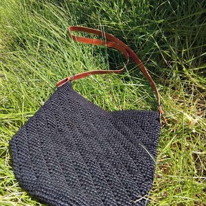 May include: A black crocheted tote bag with a brown leather strap. The bag is sitting on a bed of green grass.