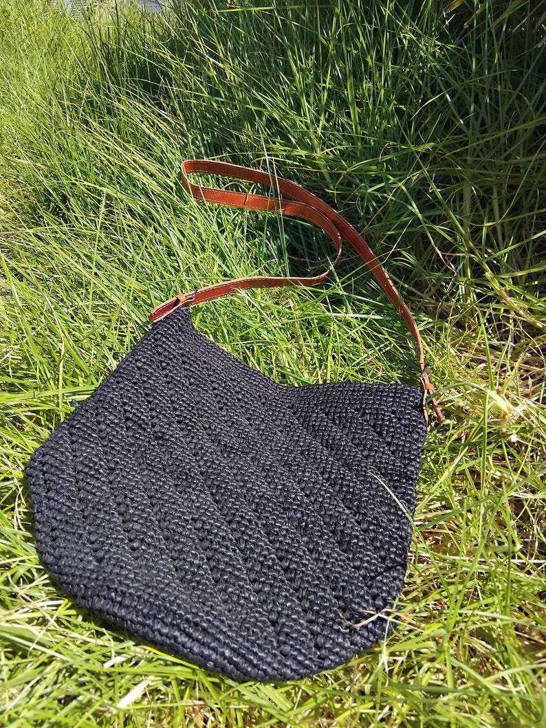 May include: A black crocheted tote bag with a brown leather strap. The bag is sitting on a bed of green grass.