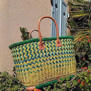 May include: A woven basket featuring a chevron pattern in shades of yellow, green, and teal. The basket has a solid green rim, a rectangular base, and two tan leather handles attached with visible stitching at the top edge.