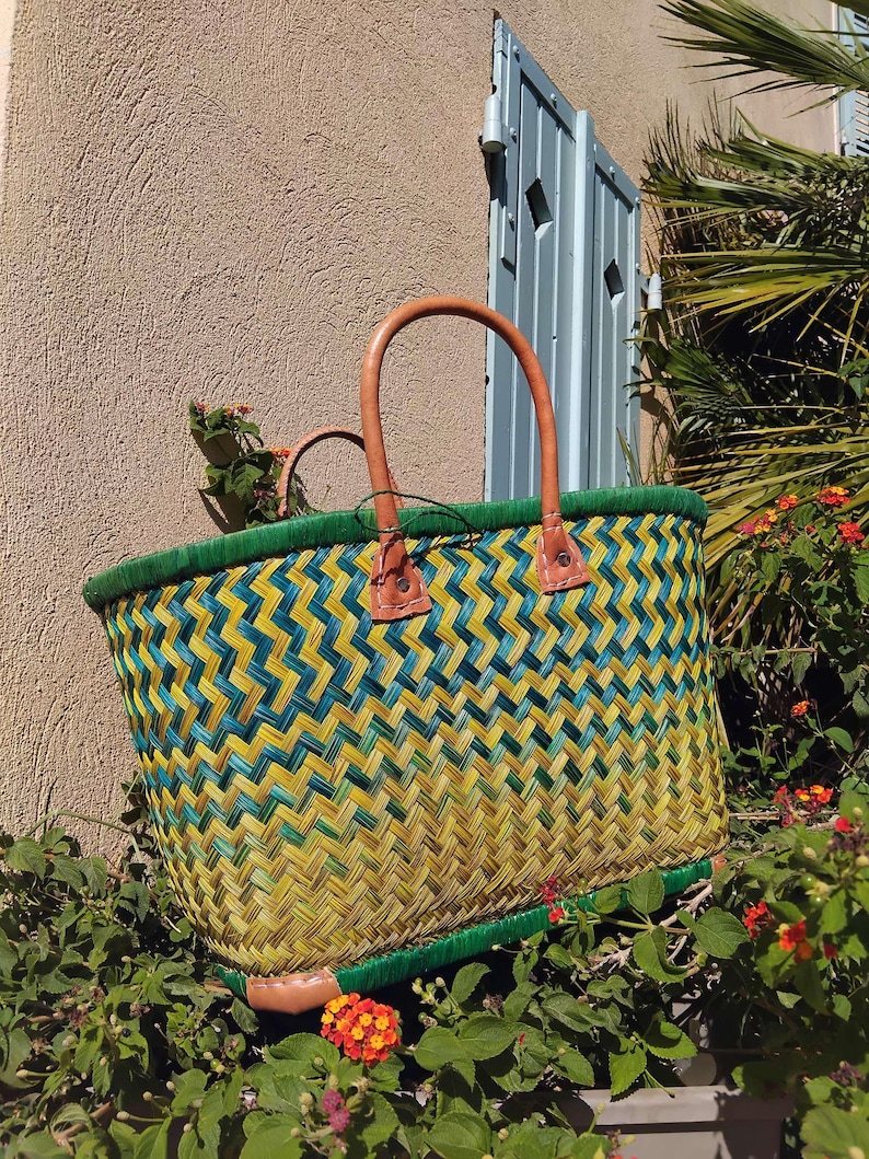 May include: A woven basket featuring a chevron pattern in shades of yellow, green, and teal. The basket has a solid green rim, a rectangular base, and two tan leather handles attached with visible stitching at the top edge.