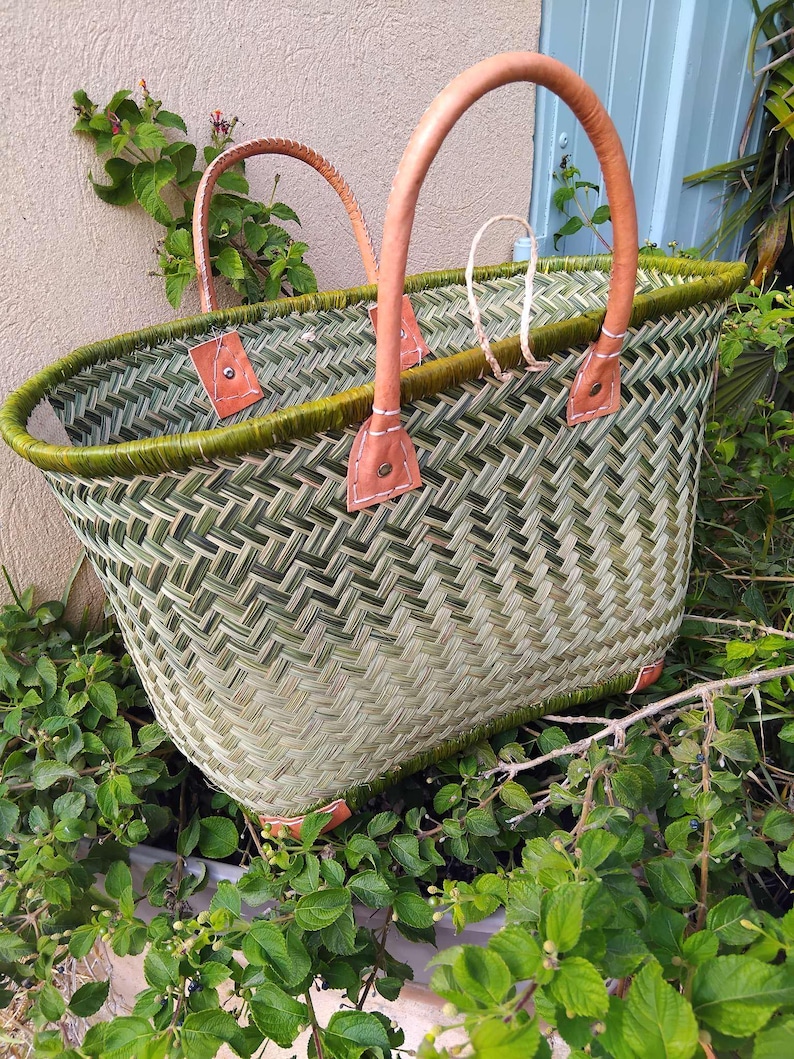 May include: A woven basket featuring a green and light tan herringbone pattern with a dark green rim. The basket includes two tan leather handles attached with metal rivets and white stitching, along with small leather corner reinforcements.