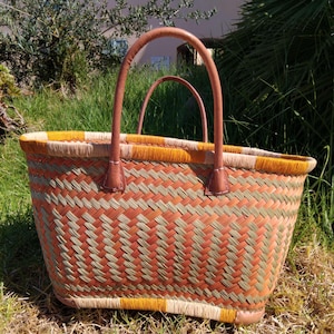 May include: A woven basket bag featuring a geometric pattern in shades of orange, olive green, and beige. The bag has two rounded tan leather handles attached with visible stitching and a reinforced base, providing a structured shape for carrying items.