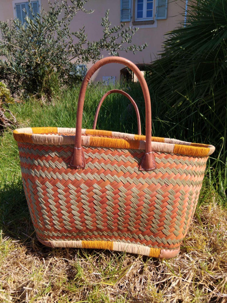 May include: A woven basket bag featuring a geometric pattern in shades of orange, olive green, and beige. The bag has two rounded tan leather handles attached with visible stitching and a reinforced base, providing a structured shape for carrying items.