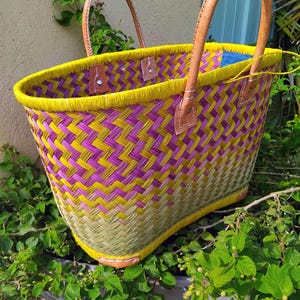May include: A woven basket bag featuring a herringbone pattern in vibrant yellow, purple, and natural green tones. The bag has a bright yellow rim and two sturdy, light brown leather handles attached with metal rivets to the woven body.