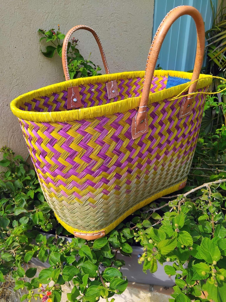 May include: A woven basket bag featuring a herringbone pattern in vibrant yellow, purple, and natural green tones. The bag has a bright yellow rim and two sturdy, light brown leather handles attached with metal rivets to the woven body.