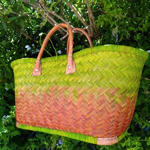 May include: A woven basket bag featuring a vibrant ombre color gradient that transitions from lime green at the top and bottom to a warm orange in the center. The bag includes two sturdy tan leather handles attached with silver metal rivets.