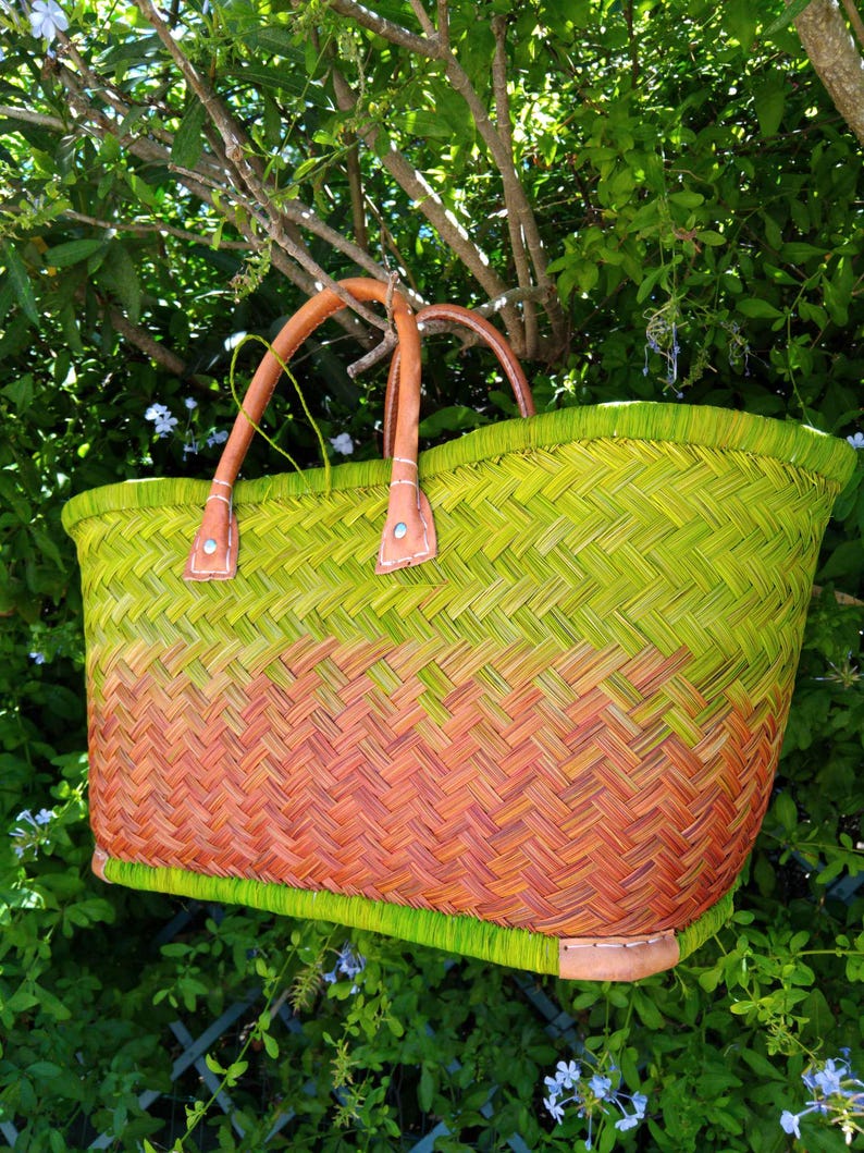 May include: A woven basket bag featuring a vibrant ombre color gradient that transitions from lime green at the top and bottom to a warm orange in the center. The bag includes two sturdy tan leather handles attached with silver metal rivets.