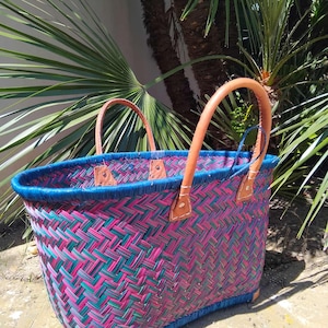 May include: A woven basket featuring a vibrant herringbone pattern in shades of magenta and teal. The basket has a solid blue rim and two tan leather handles securely attached with visible stitching. The base is reinforced with tan leather corners.