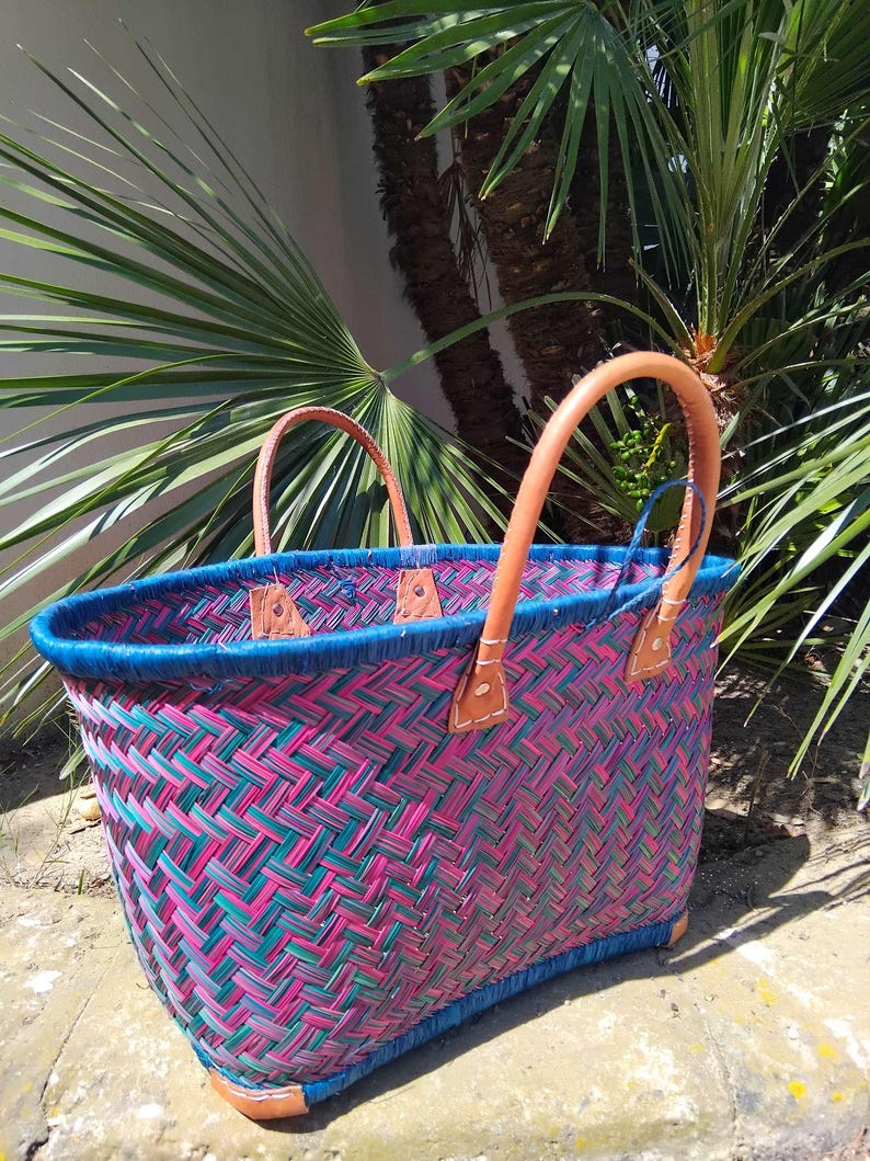 May include: A woven basket featuring a vibrant herringbone pattern in shades of magenta and teal. The basket has a solid blue rim and two tan leather handles securely attached with visible stitching. The base is reinforced with tan leather corners.