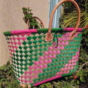 May include: A woven basket featuring a diagonal checkered pattern in vibrant pink, forest green, and natural tan fibers. The basket includes two sturdy, light brown leather handles attached with metal rivets and a reinforced pink base.