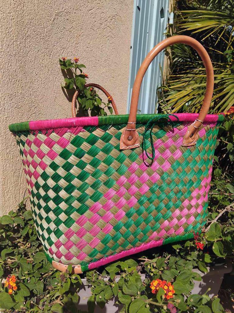 May include: A woven basket featuring a diagonal checkered pattern in vibrant pink, forest green, and natural tan fibers. The basket includes two sturdy, light brown leather handles attached with metal rivets and a reinforced pink base.