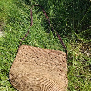 May include: A brown woven straw bag with a brown leather strap. The bag is lying on a bed of green grass.