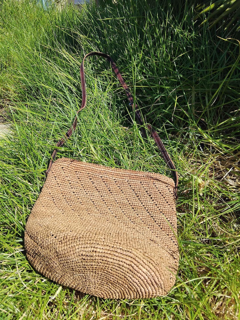 May include: A brown woven straw bag with a brown leather strap. The bag is lying on a bed of green grass.