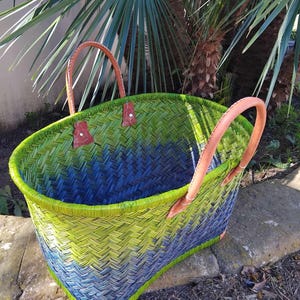 May include: A woven basket featuring a gradient pattern of lime green and deep blue. The basket has two tan leather handles attached with metal rivets and reinforced leather corners at the base, creating a structured and durable open-top design.