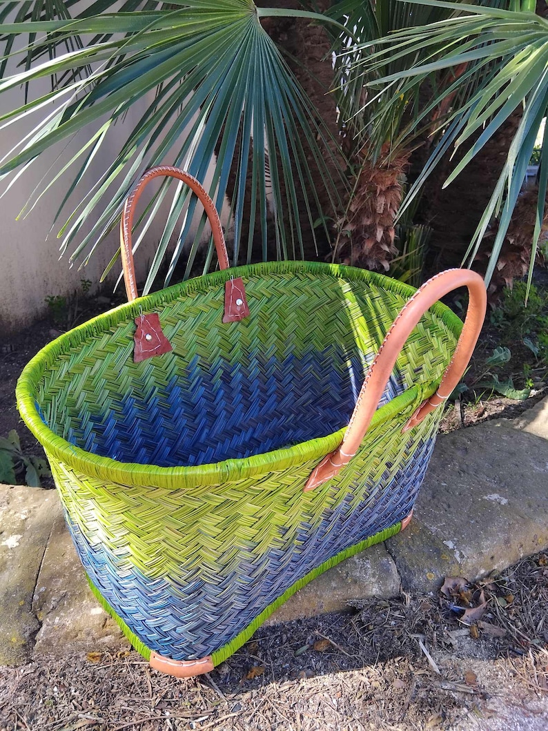 May include: A woven basket featuring a gradient pattern of lime green and deep blue. The basket has two tan leather handles attached with metal rivets and reinforced leather corners at the base, creating a structured and durable open-top design.