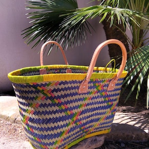 May include: A woven basket tote featuring a geometric pattern in blue, yellow, and pink with a bright yellow rim. The bag includes two light tan leather handles attached with visible stitching at the top edge of the woven body.