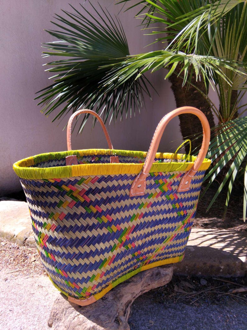 May include: A woven basket tote featuring a geometric pattern in blue, yellow, and pink with a bright yellow rim. The bag includes two light tan leather handles attached with visible stitching at the top edge of the woven body.