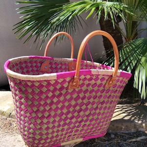 May include: A woven basket tote featuring a checkered pattern of vibrant magenta and natural tan fibers. The bag includes two sturdy, rounded tan leather handles securely attached with visible stitching and metal rivets at the top rim.