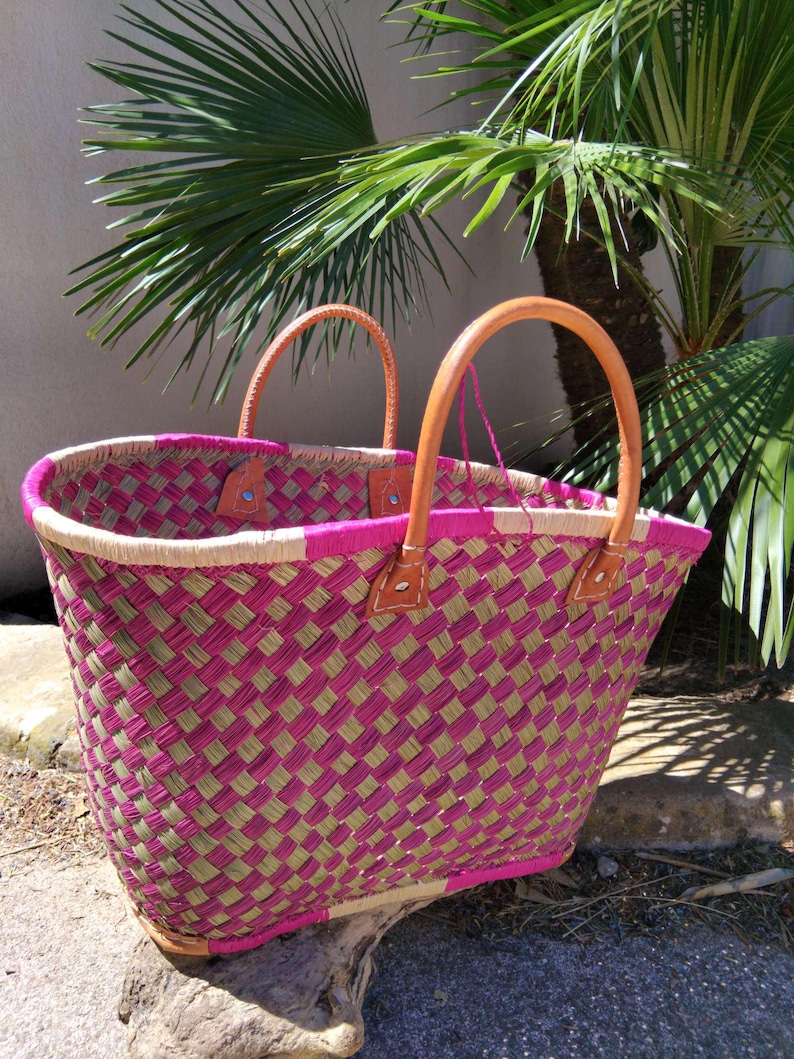 May include: A woven basket tote featuring a checkered pattern of vibrant magenta and natural tan fibers. The bag includes two sturdy, rounded tan leather handles securely attached with visible stitching and metal rivets at the top rim.