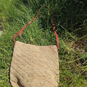 May include: A straw tote bag with a brown leather strap. The bag is a light brown colour and has a woven texture. The bag is sitting on a bed of green grass.