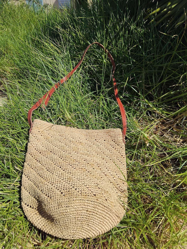May include: A straw tote bag with a brown leather strap. The bag is a light brown colour and has a woven texture. The bag is sitting on a bed of green grass.