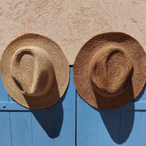May include: Two woven straw hats with wide brims. One hat is a light tan color, and the other is a darker brown. The hats are hanging on a blue surface against a textured beige wall. The hats are circular with a spiral pattern.