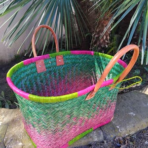 May include: A woven basket featuring a herringbone pattern in vibrant shades of green and pink. The basket has a bright lime green rim, two tan leather handles secured with metal rivets, and tan leather corner reinforcements at the base.