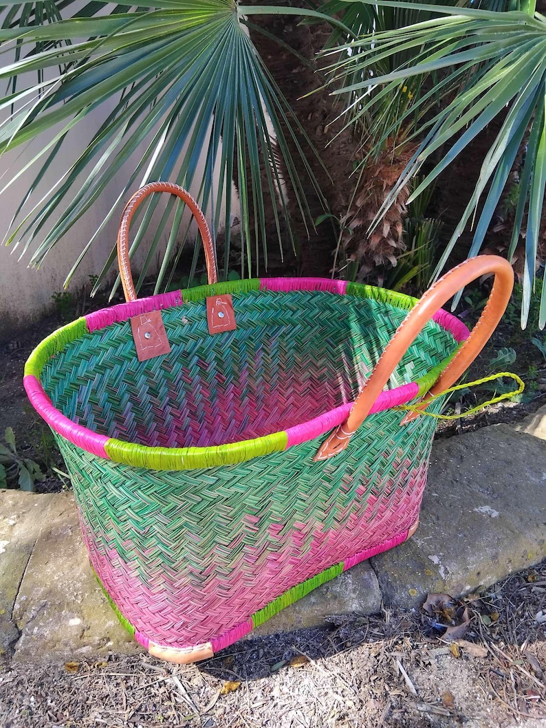 May include: A woven basket featuring a herringbone pattern in vibrant shades of green and pink. The basket has a bright lime green rim, two tan leather handles secured with metal rivets, and tan leather corner reinforcements at the base.
