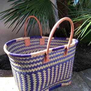 May include: A woven basket with a chevron pattern in purple and natural tan fibers. The basket features two tan leather handles attached with white stitching and small tan leather patches at the top rim and base corners for reinforcement.