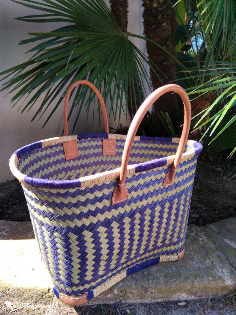 May include: A woven basket with a chevron pattern in purple and natural tan fibers. The basket features two tan leather handles attached with white stitching and small tan leather patches at the top rim and base corners for reinforcement.