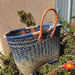 May include: A woven basket with a herringbone pattern in shades of blue and natural tan. The basket features two tan leather handles attached with metal rivets and a blue woven rim. The structure is open-topped with a reinforced base.