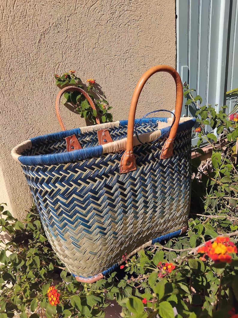 May include: A woven basket with a herringbone pattern in shades of blue and natural tan. The basket features two tan leather handles attached with metal rivets and a blue woven rim. The structure is open-topped with a reinforced base.