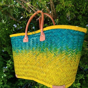 May include: A woven basket tote bag featuring a vibrant ombre color gradient that transitions from a deep teal at the top to a bright yellow at the base. The bag includes two tan leather handles and leather reinforcement patches.