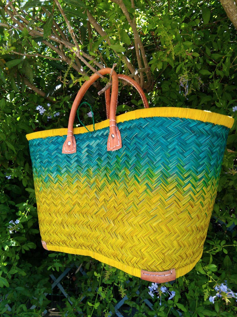 May include: A woven basket tote bag featuring a vibrant ombre color gradient that transitions from a deep teal at the top to a bright yellow at the base. The bag includes two tan leather handles and leather reinforcement patches.