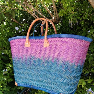 May include: A woven basket bag featuring a gradient pattern that transitions from a deep teal at the base to a vibrant magenta at the top. The bag includes a blue rim, two tan leather handles secured with white stitching, and tan leather corner accents.