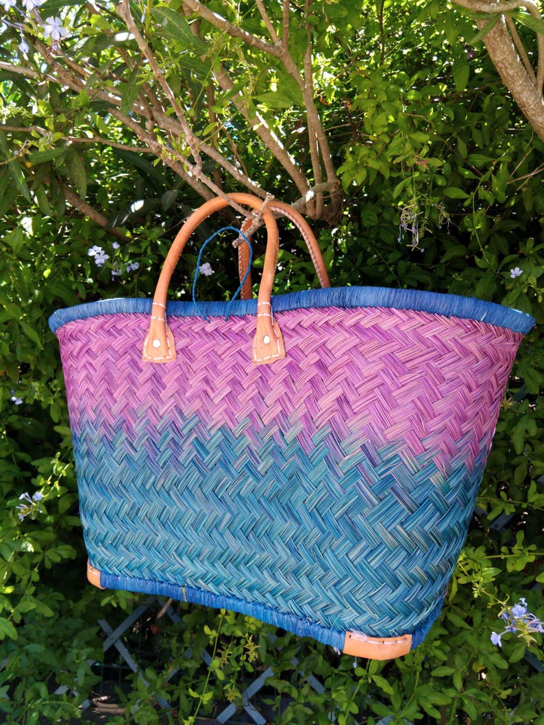 May include: A woven basket bag featuring a gradient pattern that transitions from a deep teal at the base to a vibrant magenta at the top. The bag includes a blue rim, two tan leather handles secured with white stitching, and tan leather corner accents.