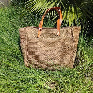 May include: A rectangular tote bag crafted from woven tan straw material. The bag features two adjustable brown leather handles attached with silver-toned metal buckles and rivets. The structure is defined by vertical side panels and a flat base.