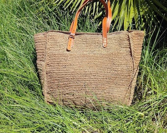 May include: A rectangular tote bag crafted from woven tan straw material. The bag features two adjustable brown leather handles attached with silver-toned metal buckles and rivets. The structure is defined by vertical side panels and a flat base.