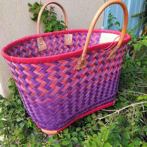 May include: A woven basket featuring a vibrant herringbone pattern in shades of purple and red. The basket has a bright red rim and two tan leather handles secured with metal rivets. The structure is open-topped with a rectangular base.
