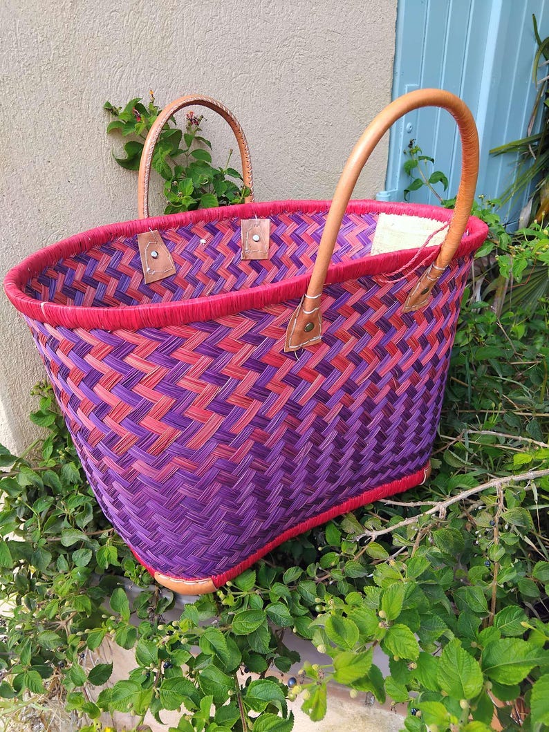 May include: A woven basket featuring a vibrant herringbone pattern in shades of purple and red. The basket has a bright red rim and two tan leather handles secured with metal rivets. The structure is open-topped with a rectangular base.