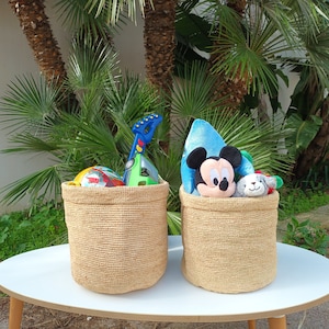 Peut inclure: Deux paniers en osier naturel remplis de jouets. Le panier à gauche contient une guitare jouet, une voiture jouet et un ballon. Le panier à droite contient un Mickey Mouse en peluche, un animal en peluche et un jouet bleu et vert. Les paniers sont posés sur une table blanche.
