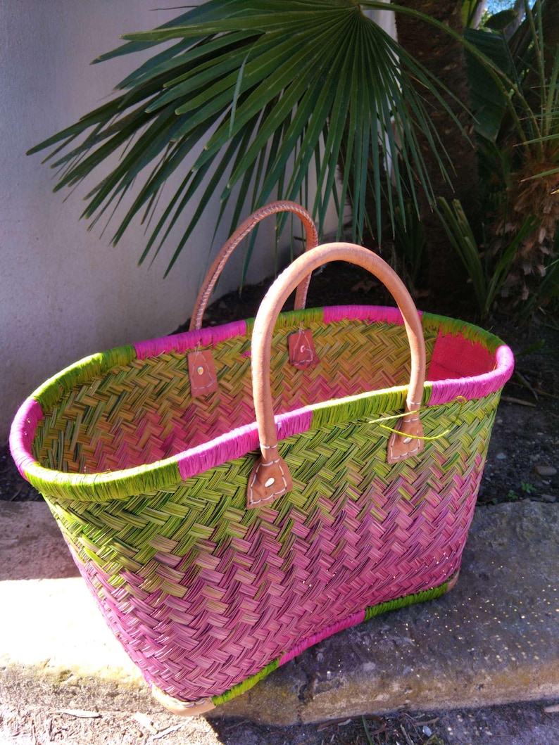 May include: A woven basket featuring a vibrant herringbone pattern in shades of bright pink and lime green. The basket includes two sturdy, light tan leather handles securely attached with visible stitching to the top rim.