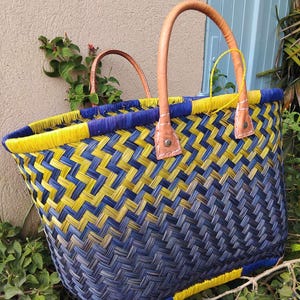 May include: A woven basket tote featuring a diagonal ombre pattern transitioning from bright yellow to deep navy blue. The bag includes two tan leather handles secured with metal rivets and a yellow woven rim, showcasing a structured, open-top design.