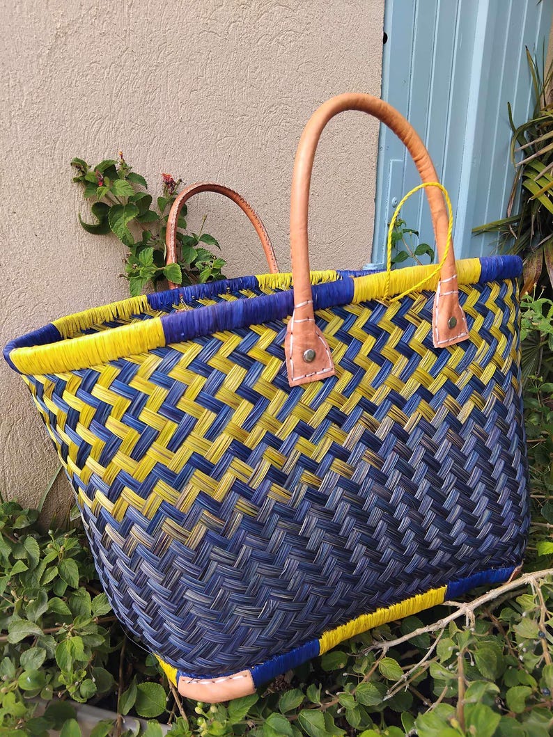 May include: A woven basket tote featuring a diagonal ombre pattern transitioning from bright yellow to deep navy blue. The bag includes two tan leather handles secured with metal rivets and a yellow woven rim, showcasing a structured, open-top design.