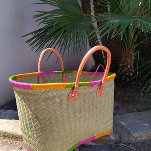 May include: A woven straw basket featuring two tan leather handles attached with visible stitching. The top and bottom edges are decorated with alternating sections of bright pink, lime green, and orange thread wrapped around the rim.