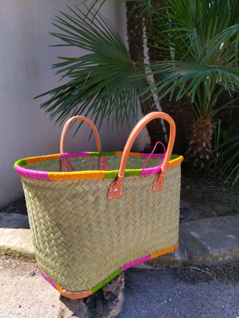 May include: A woven straw basket featuring two tan leather handles attached with visible stitching. The top and bottom edges are decorated with alternating sections of bright pink, lime green, and orange thread wrapped around the rim.