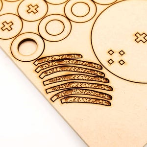May include: A close-up of a wooden sheet with laser-cut shapes. The shapes include circles, plus signs, and curved lines with dotted patterns. The wood has a light brown color and a smooth texture. The design suggests a craft or DIY project.